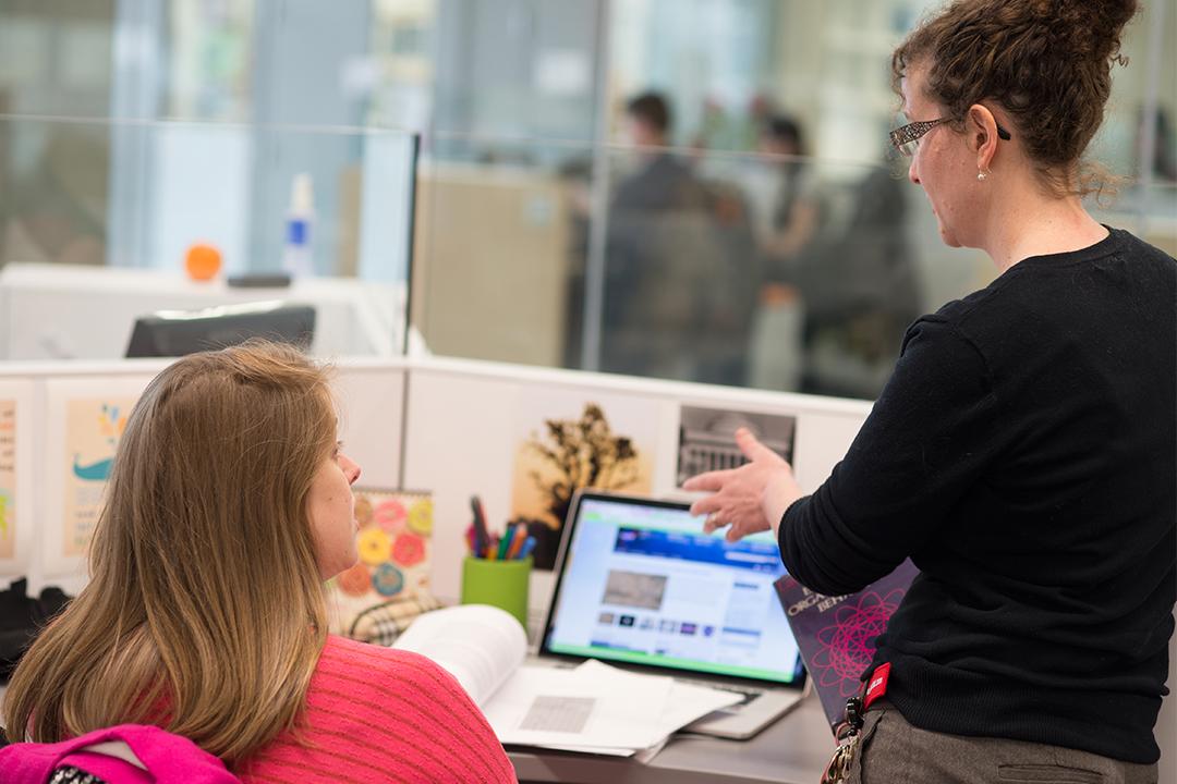 Student sitting at desk with laptop open, teacher standing and talking to her